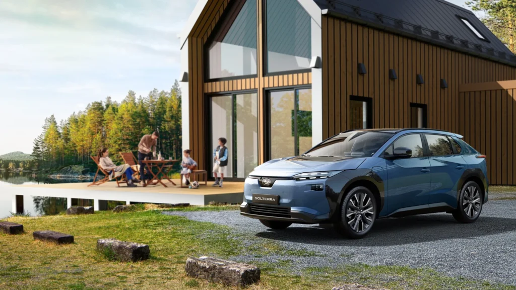 Blå Subaru Solterra parkert utenfor et hus ved innsjøen med folk på dekk.