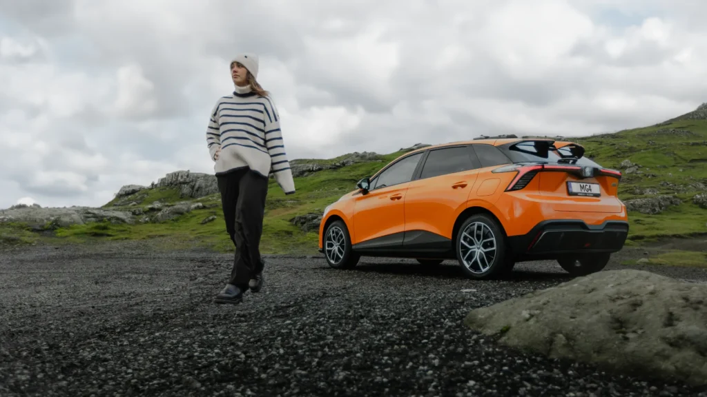 Person går nær orange bil i steinete, gressrike landskap.