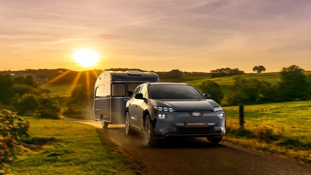 Blå SUV trekker campingvogn på landevei i solnedgang.