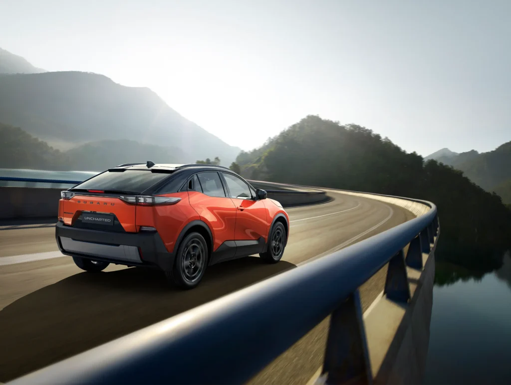 Oransje SUV kjører på svingete fjellvei ved vann under klar himmel.