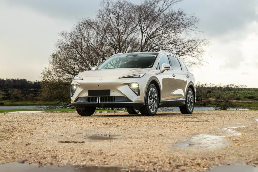 Beige elbil SUV parkert ute på sand, tre og skyer bak.