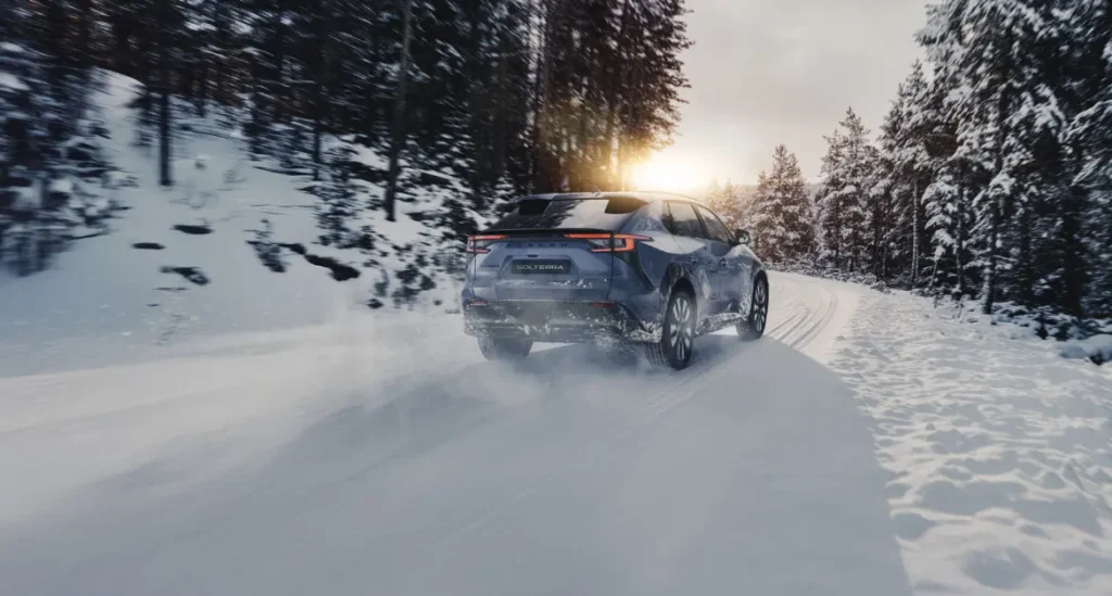 Blå Subaru Solterra kjører på snødekt vei gjennom skog.
