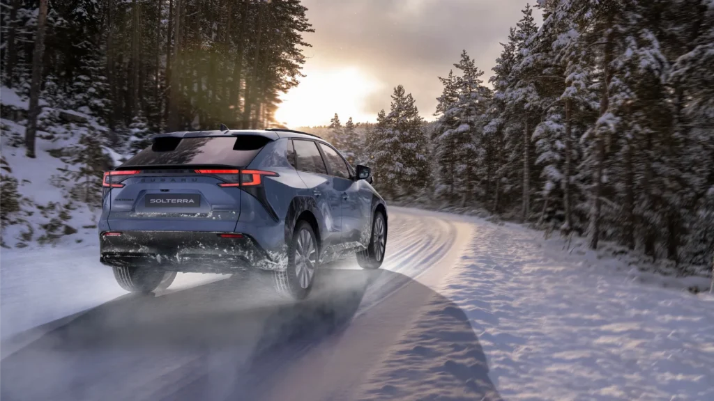 Blå Subaru Solterra kjører på snødekt, svingete vei i solnedgang.