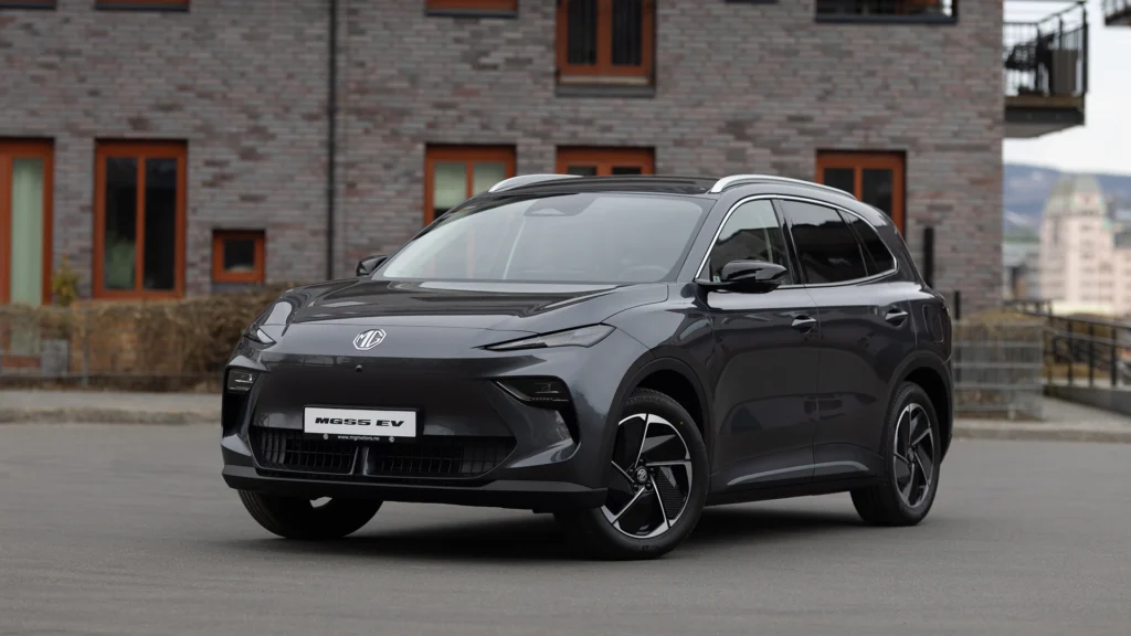 Mørk grå elbil utendørs foran moderne murbygning med store vinduer.